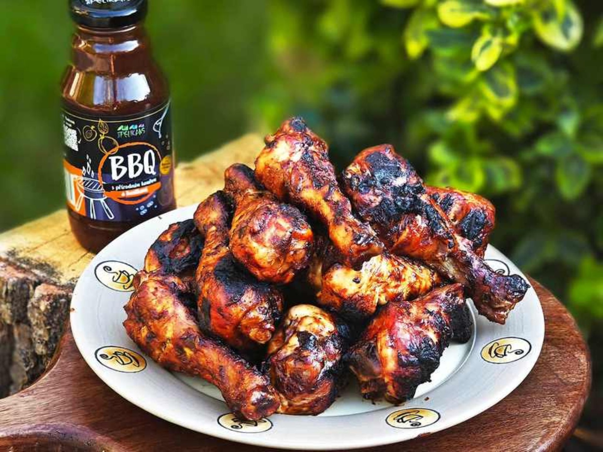 Grilovaná kuřecí křídla na talíři s omáčkou BBQ v lahvičce, venkovní zelené pozadí.
